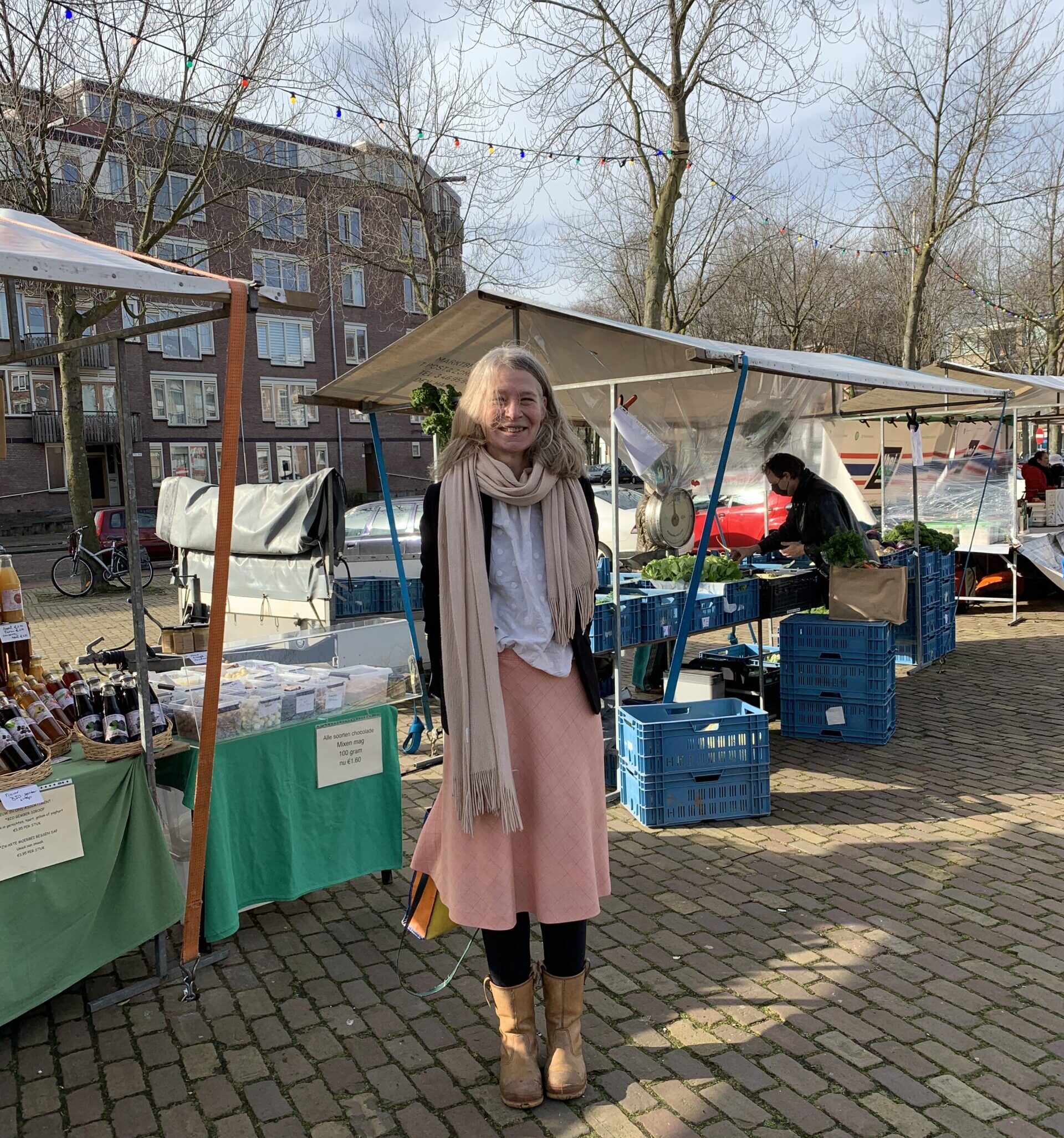 Woman wearing a pink skirt, a black jacket and a scarf is standing in front of market stalls