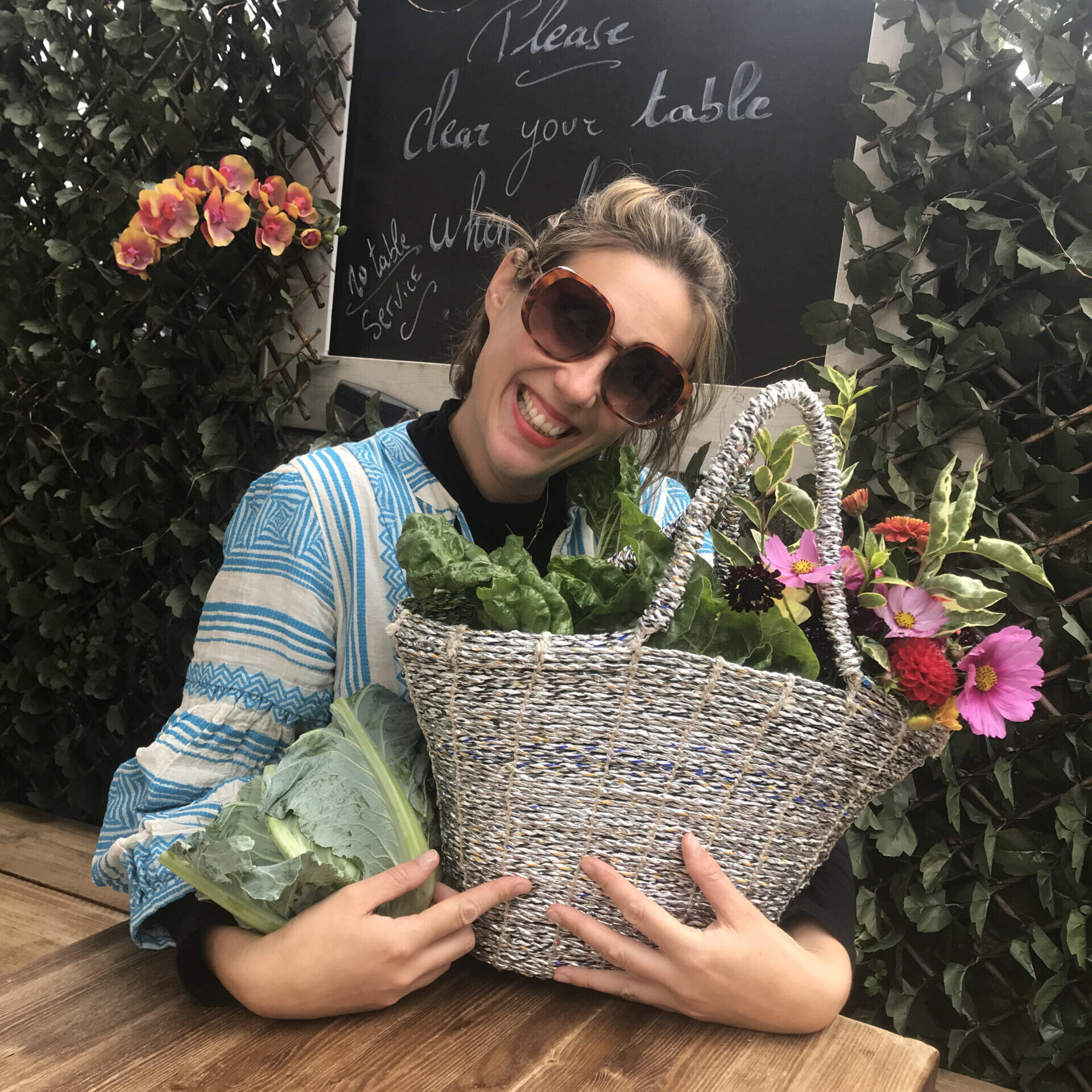 woman in blue dress and sunglasses sitting at a table holding a cauliflower and a basket filled with flowers