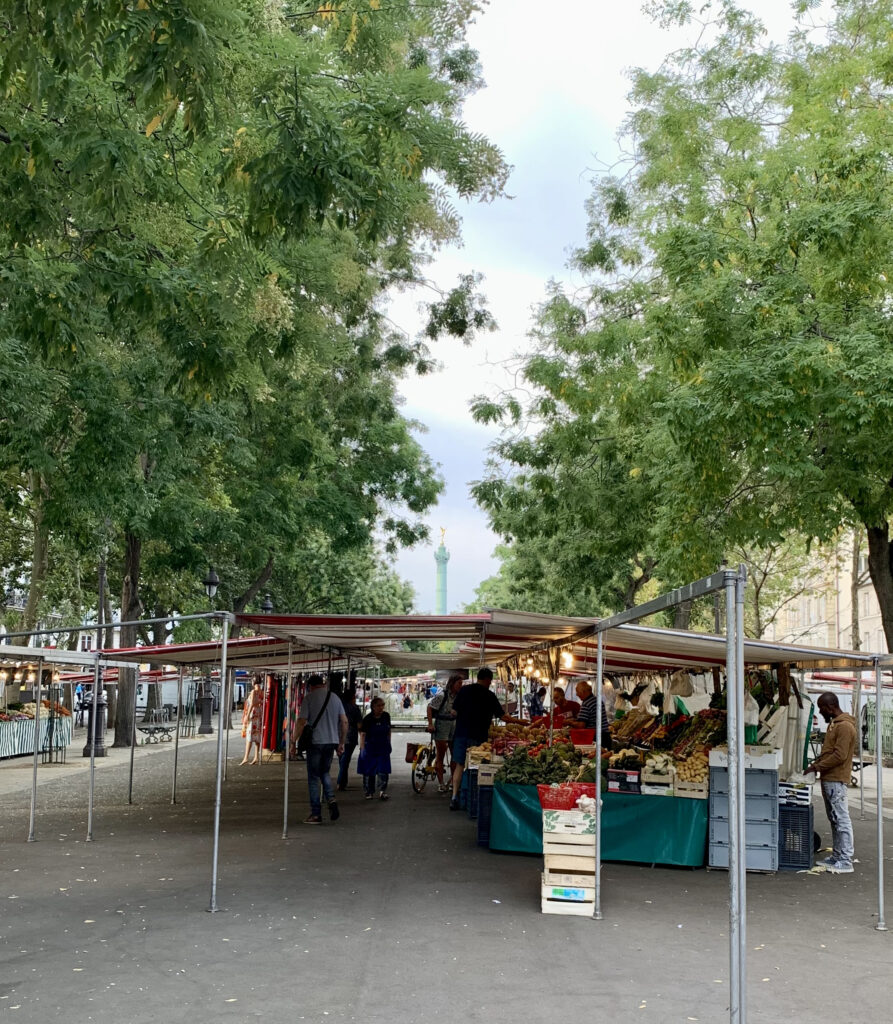 a street with market stalls under trees in daylight with the July Column in Paris in the background