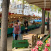 A bustling Paris outdoor market with vendor stalls selling fresh produce. In the foreground, wooden crates filled with fruits and vegetables sit alongside bunches of pink radishes. Shoppers browse the green-covered stalls under strings of lights, with classic Parisian buildings visible in the background.