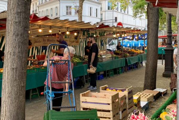 A bustling Paris outdoor market with vendor stalls selling fresh produce. In the foreground, wooden crates filled with fruits and vegetables sit alongside bunches of pink radishes. Shoppers browse the green-covered stalls under strings of lights, with classic Parisian buildings visible in the background.