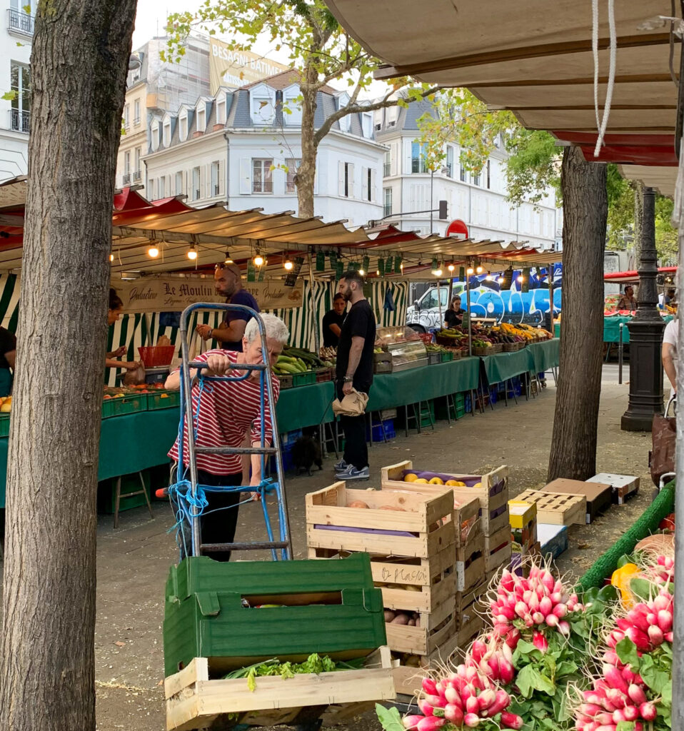 A bustling Paris outdoor market with vendor stalls selling fresh produce. In the foreground, wooden crates filled with fruits and vegetables sit alongside bunches of pink radishes. Shoppers browse the green-covered stalls under strings of lights, with classic Parisian buildings visible in the background.