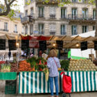 lady standing in front of market stall full of produce with a red shopping bag - Markets in Paris Today