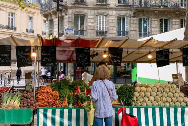 lady standing in front of market stall full of produce with a red shopping bag - Markets in Paris Today