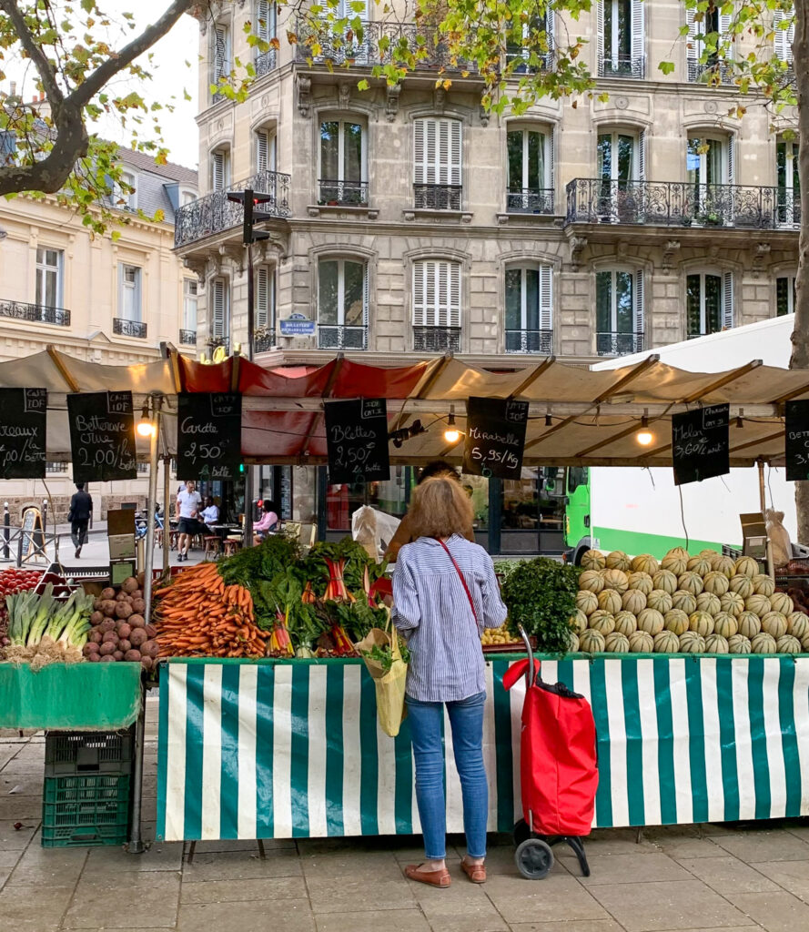 lady standing in front of market stall full of produce with a red shopping bag - Markets in Paris Today