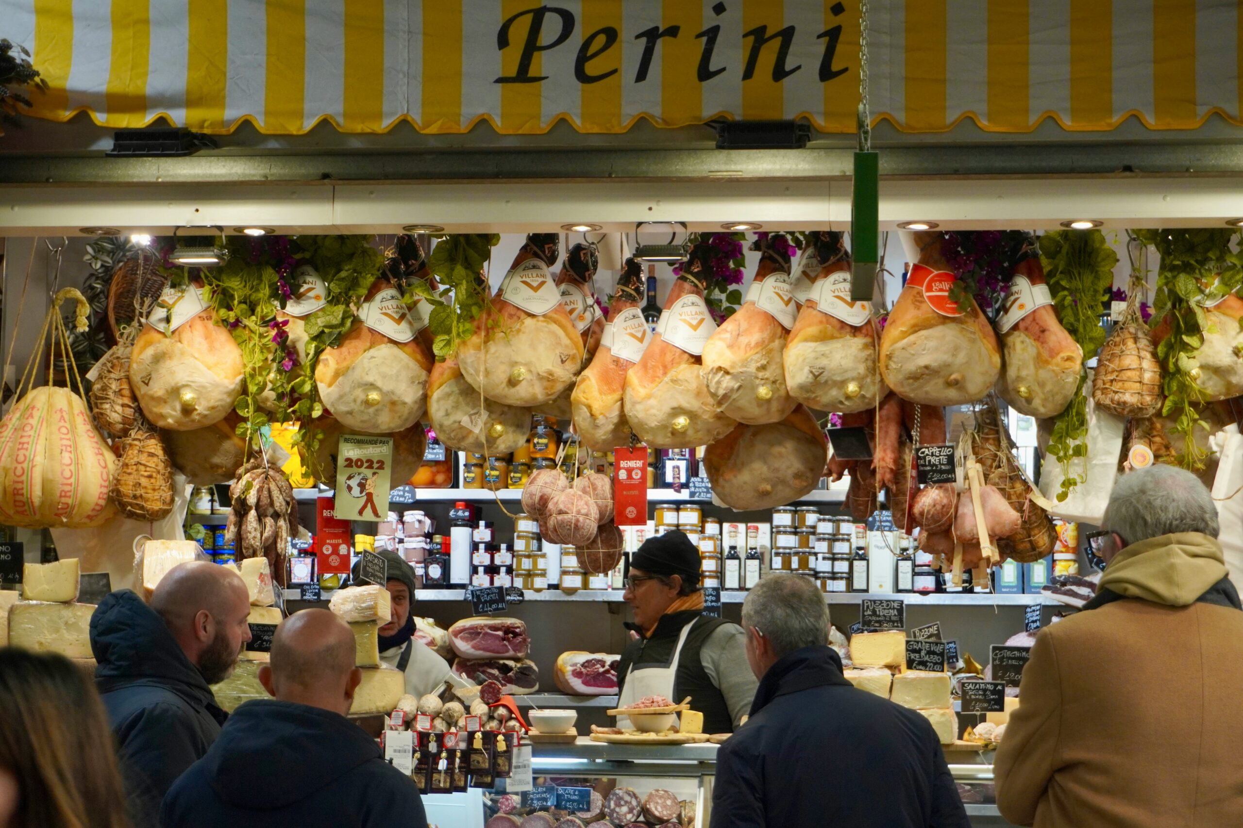 Italian deli 'Perini' with yellow striped awning. Hanging prosciutto legs and cured meats above a counter displaying cheeses and specialty foods. Vendors in white aprons serving customers.