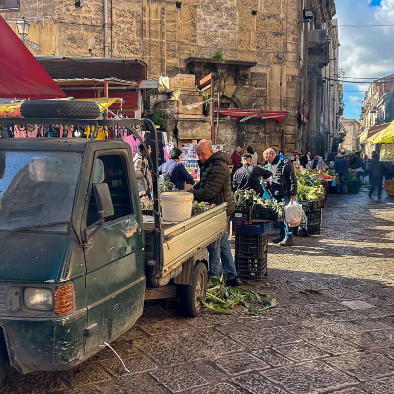 Palermo guide Bustling market street in Palermo with a small green delivery truck parked as vendors unload fresh vegetables while shoppers browse produce stands beneath colourful awnings along ancient stone buildings