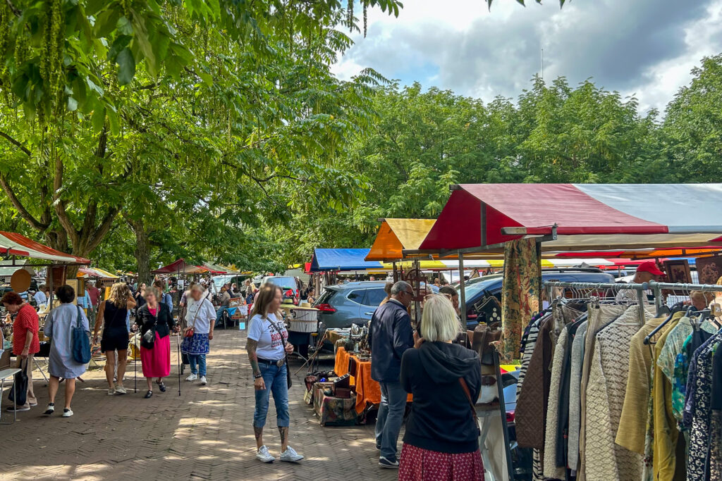 Amstelveld Antique Market Amsterdam - Alt text: Outdoor market with colorful stall canopies under green trees. Shoppers browse clothing racks and merchandise on a brick pathway. People walking between vendor stalls on a sunny day with some clouds overhead.