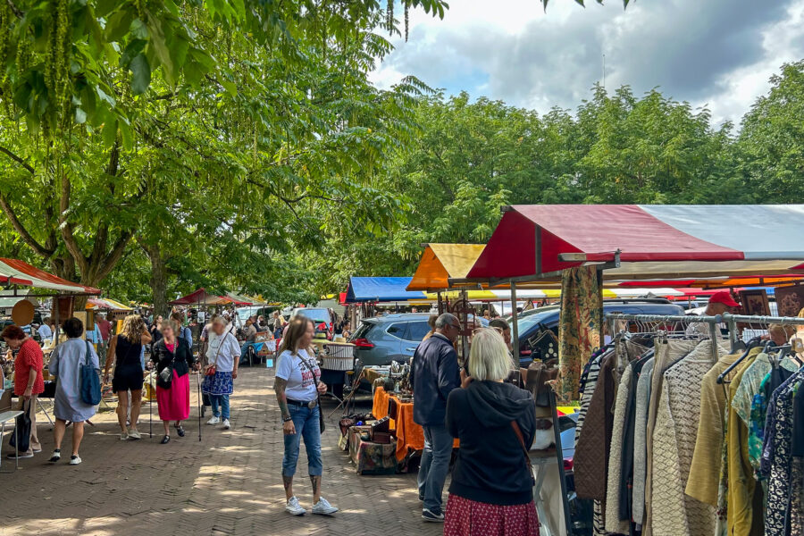 Amstelveld Antique Market Amsterdam - Alt text: Outdoor market with colorful stall canopies under green trees. Shoppers browse clothing racks and merchandise on a brick pathway. People walking between vendor stalls on a sunny day with some clouds overhead.