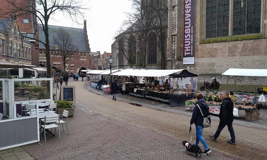 Kerkplein Cheese Market Alkmaar - Market stalls with white canopies along a cobblestone street in Alkmaar, next to a large church. Two people walk a small dog past outdoor café seating and vendor stalls selling flowers and goods on a cloudy day. Traditional Dutch brick buildings frame the scene