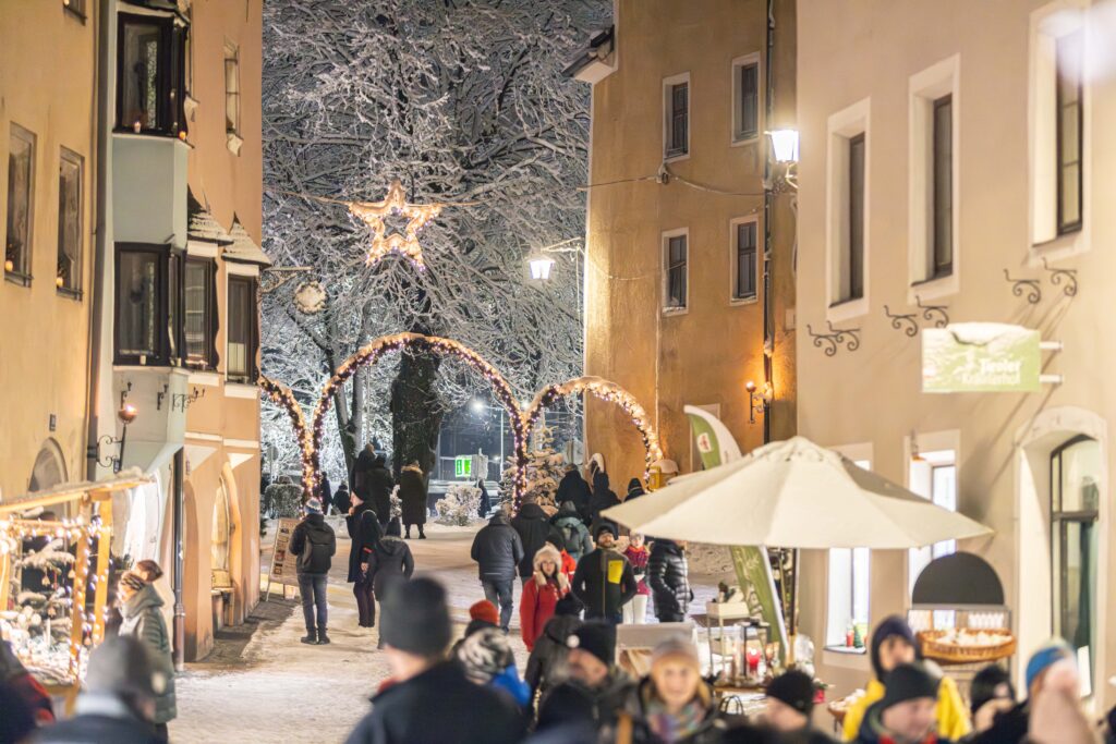 Snowy evening at Rattenberg Christmas Market. Visitors walk through a narrow medieval street with illuminated arches, a star decoration in snow-covered trees, and market stalls beneath golden-lit historic buildings.