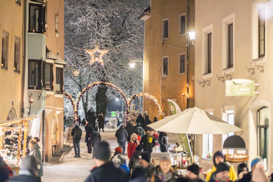 Snowy evening at Rattenberg Christmas Market. Visitors walk through a narrow medieval street with illuminated arches, a star decoration in snow-covered trees, and market stalls beneath golden-lit historic buildings.