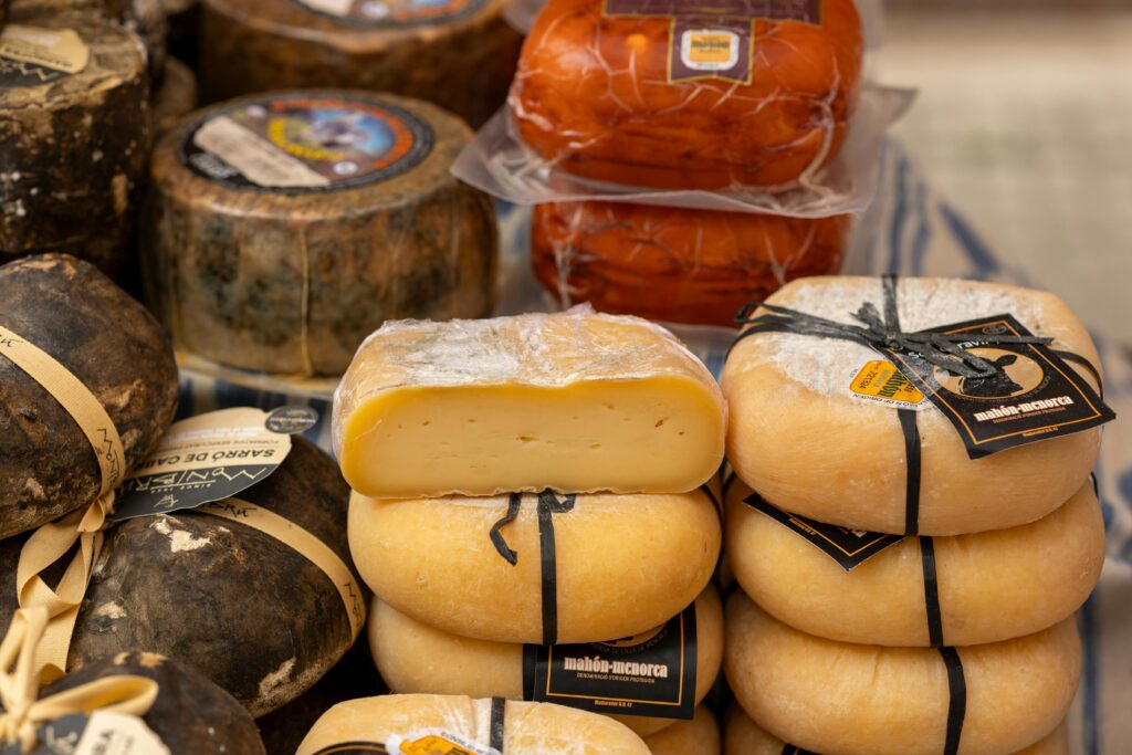 Santa Maria Market Mallorca - Artisanal cheeses on display including stacked yellow Mahón-Menorca wheels, aged dark cheeses, and orange cheese in packaging.