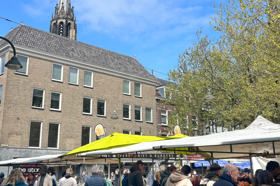 Delft market: A bustling outdoor market scene in Delft, Netherlands on a sunny day. Crowds of people browse market stalls with white and bright yellow canopies selling flowers and food. In the background, the distinctive Gothic spire of the Nieuwe Kerk (New Church) rises against a blue sky with scattered clouds. A brick building with a traditional Dutch gabled roof stands between the market and the church. Fresh spring trees with bright green leaves frame the right side of the image. Signs visible include "Verse Friet & Burgers" (Fresh Fries & Burgers) on one of the food stalls.