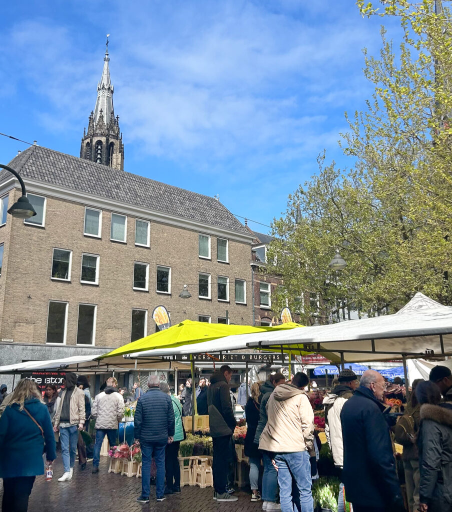 Delft market: A bustling outdoor market scene in Delft, Netherlands on a sunny day. Crowds of people browse market stalls with white and bright yellow canopies selling flowers and food. In the background, the distinctive Gothic spire of the Nieuwe Kerk (New Church) rises against a blue sky with scattered clouds. A brick building with a traditional Dutch gabled roof stands between the market and the church. Fresh spring trees with bright green leaves frame the right side of the image. Signs visible include "Verse Friet & Burgers" (Fresh Fries & Burgers) on one of the food stalls.