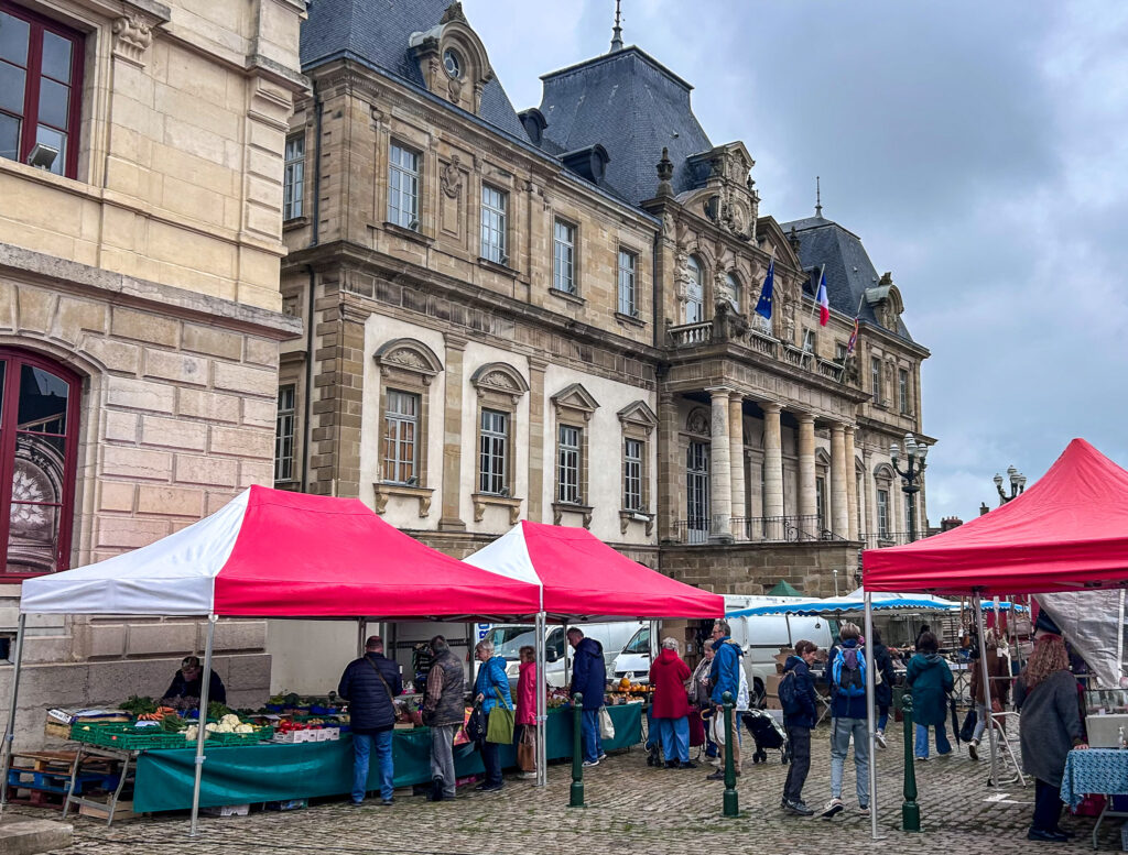 Autun market with red and white vendor tents in front of the town hall, showing fresh produce stalls and shoppers on a cloudy day in Burgundy, France.