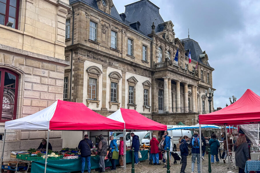 Autun market with red and white vendor tents in front of the town hall, showing fresh produce stalls and shoppers on a cloudy day in Burgundy, France.