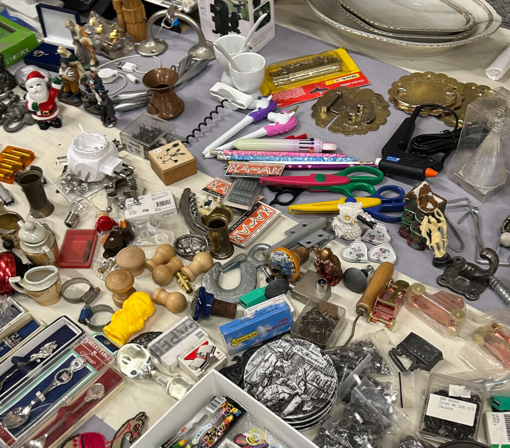 Vijfhuizen flea market: A flea market table filled with a mix of secondhand items including scissors, colourful pens, wooden stamps, small figurines, brass hardware, ceramic heart-shaped trinkets, serving plates, a copper coffee pot, decorative tiles, office supplies, and various other odds and ends.