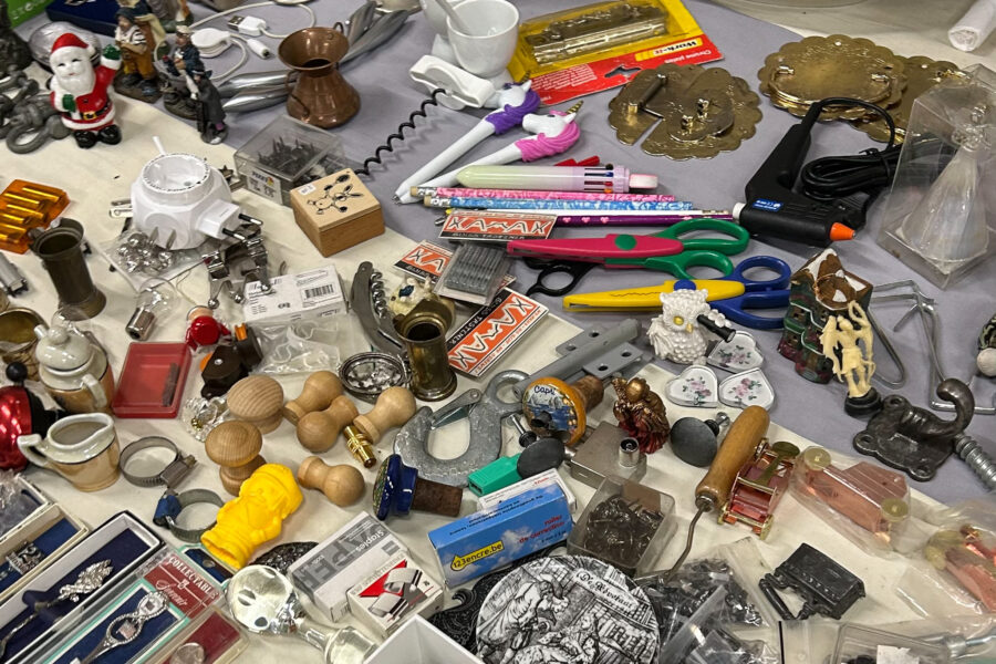 Vijfhuizen flea market: A flea market table filled with a mix of secondhand items including scissors, colourful pens, wooden stamps, small figurines, brass hardware, ceramic heart-shaped trinkets, serving plates, a copper coffee pot, decorative tiles, office supplies, and various other odds and ends.