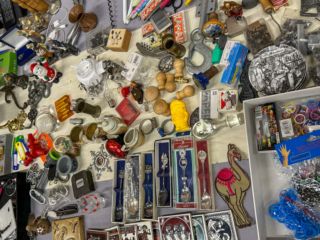 Eemnes Flea Market. A flea market table covered with a wide mix of small items, including collectible souvenir spoons in display cases, miniature figurines, brass ornaments, wooden stamps, a corkscrew, loom bands, a camel-shaped patch, decorative tiles, keychains, and various other knick-knacks and bric-a-brac.