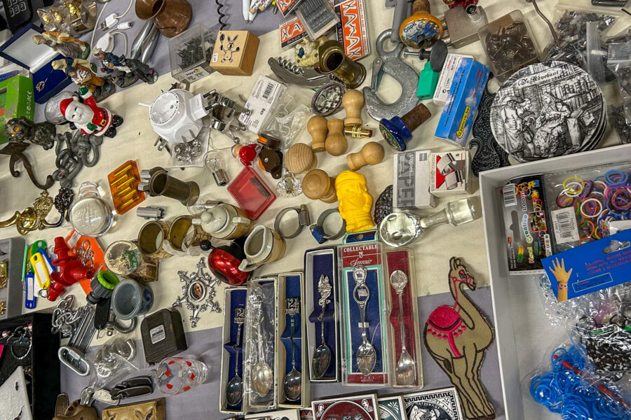 Eemnes Flea Market. A flea market table covered with a wide mix of small items, including collectible souvenir spoons in display cases, miniature figurines, brass ornaments, wooden stamps, a corkscrew, loom bands, a camel-shaped patch, decorative tiles, keychains, and various other knick-knacks and bric-a-brac.