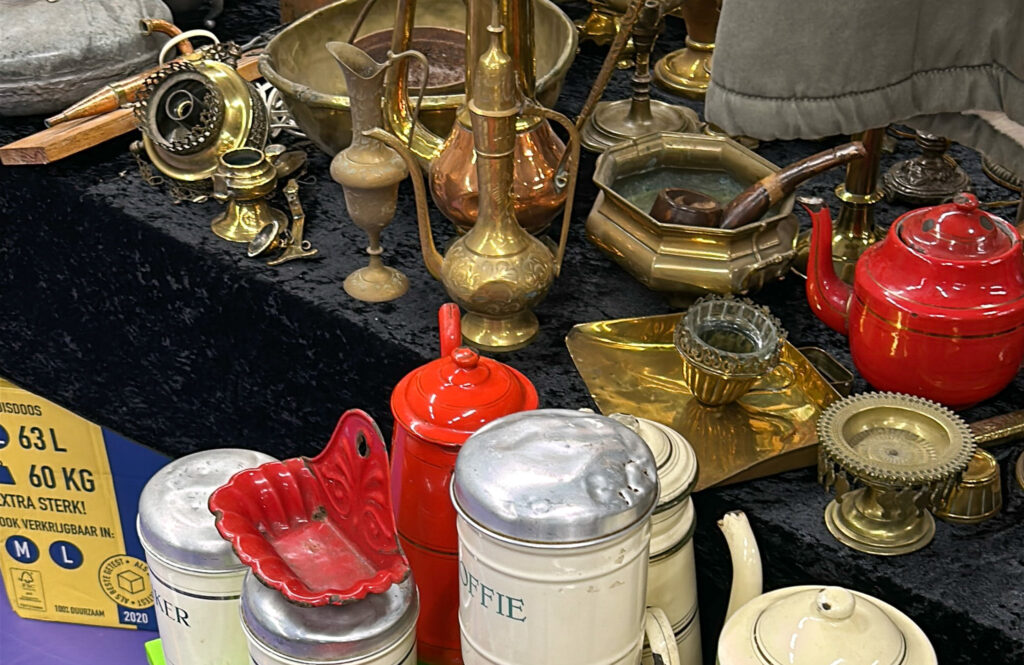 Zeist Flea Market: A flea market stall displaying vintage brass and copper items including pitchers, candlesticks, and an ashtray, alongside red enamel kitchenware, old Dutch coffee and sugar canisters, a small teapot, and a glass candle holder, all laid out on a dark cloth