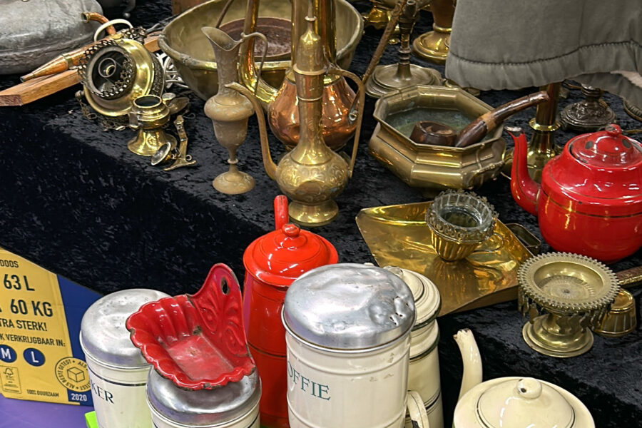 Zeist Flea Market: A flea market stall displaying vintage brass and copper items including pitchers, candlesticks, and an ashtray, alongside red enamel kitchenware, old Dutch coffee and sugar canisters, a small teapot, and a glass candle holder, all laid out on a dark cloth